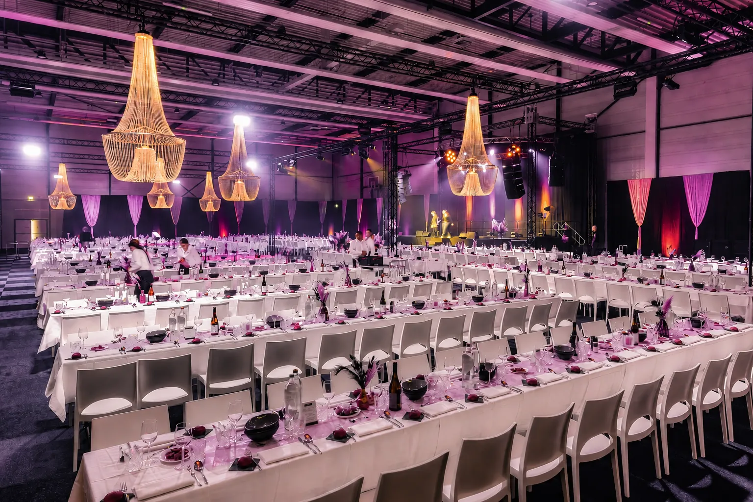 Volledig ingerichte evenementenlocatie met lange dinertafels, sfeervolle verlichting en podium voor een zakelijk evenement.